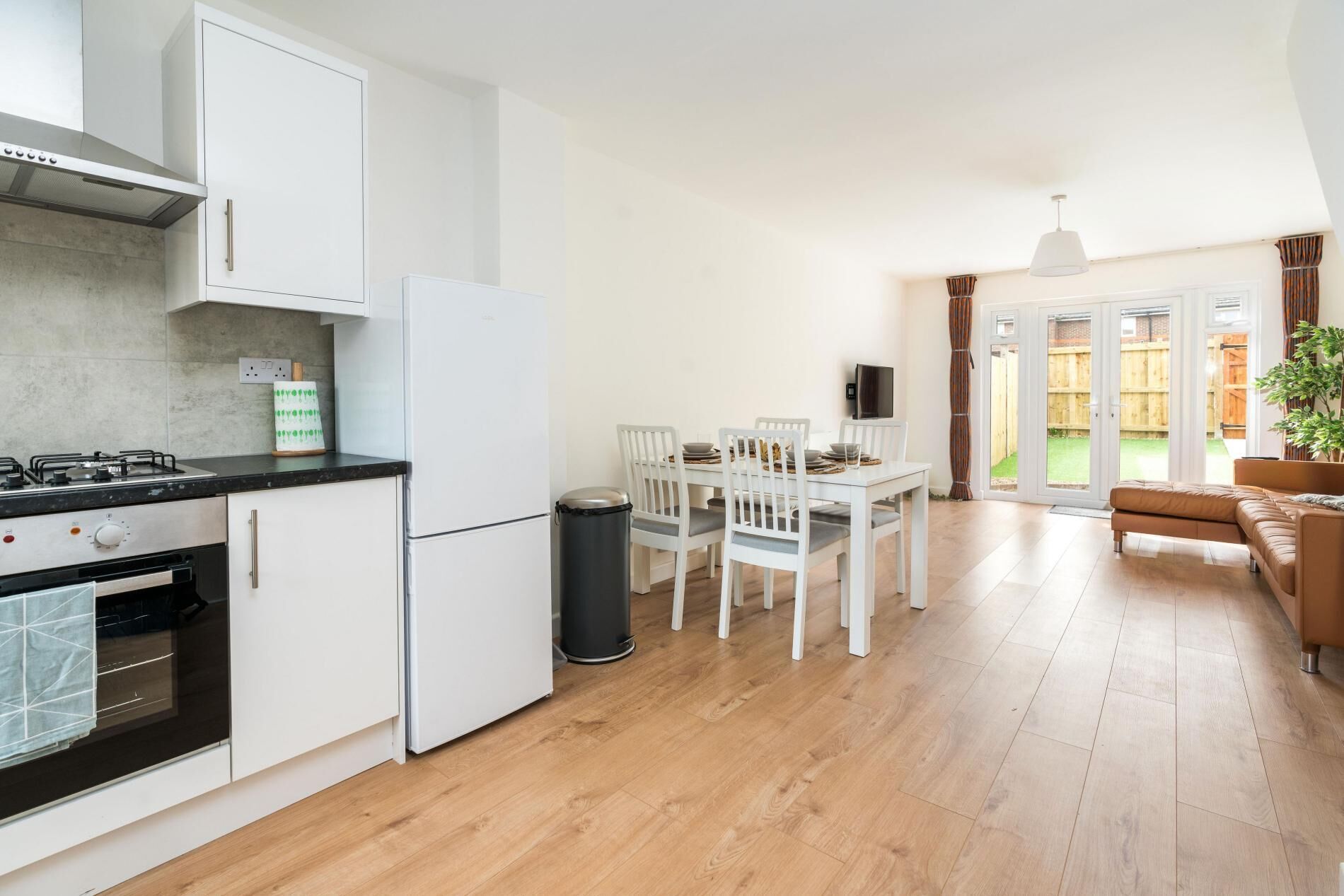 A modern kitchen with white cabinets, a gas stove, and a refrigerator. There is a dining area with a table and chairs, and a living room with a sofa and a TV.