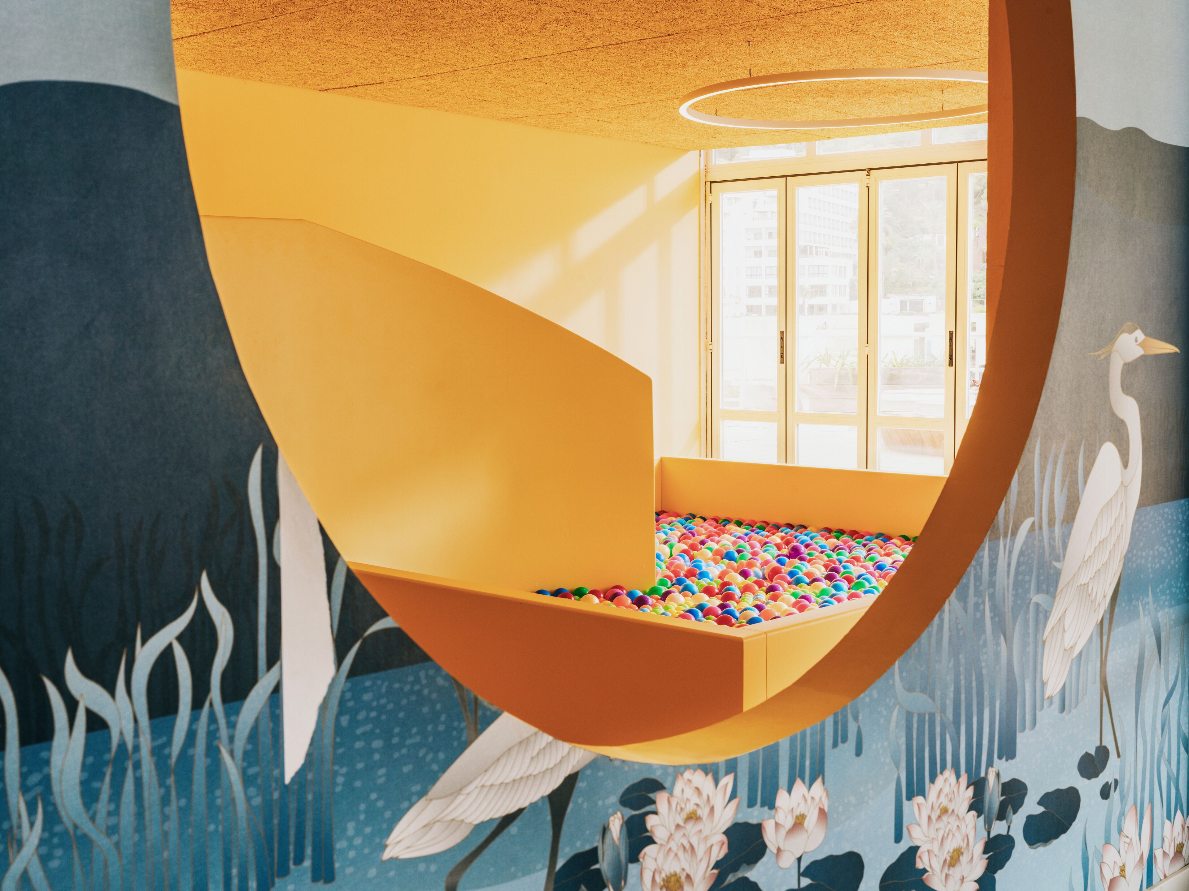 A room with a colorful ball pit, a window with a view of buildings, and a mural with birds and lotus flowers.