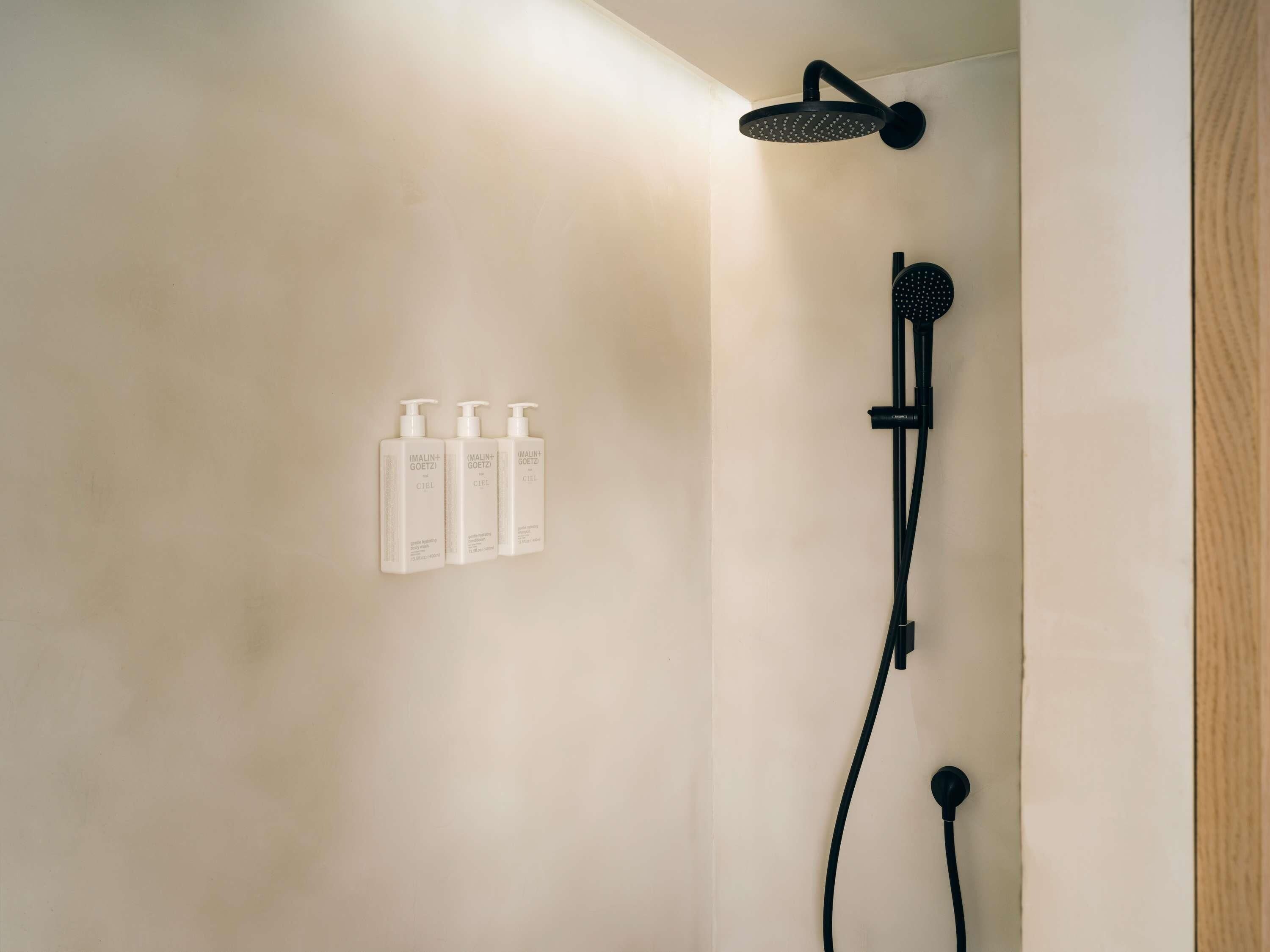 A shower area with a handheld showerhead and a wall-mounted faucet. There are three bottles of shampoo on the wall.