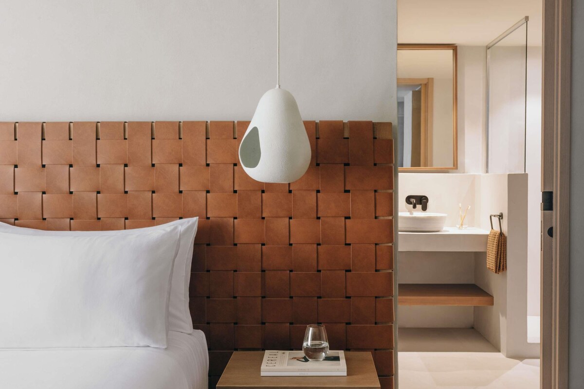 A hotel room with a bed, a bedside table with a book and a glass, a hanging light fixture, a bathroom with a mirror and sink.