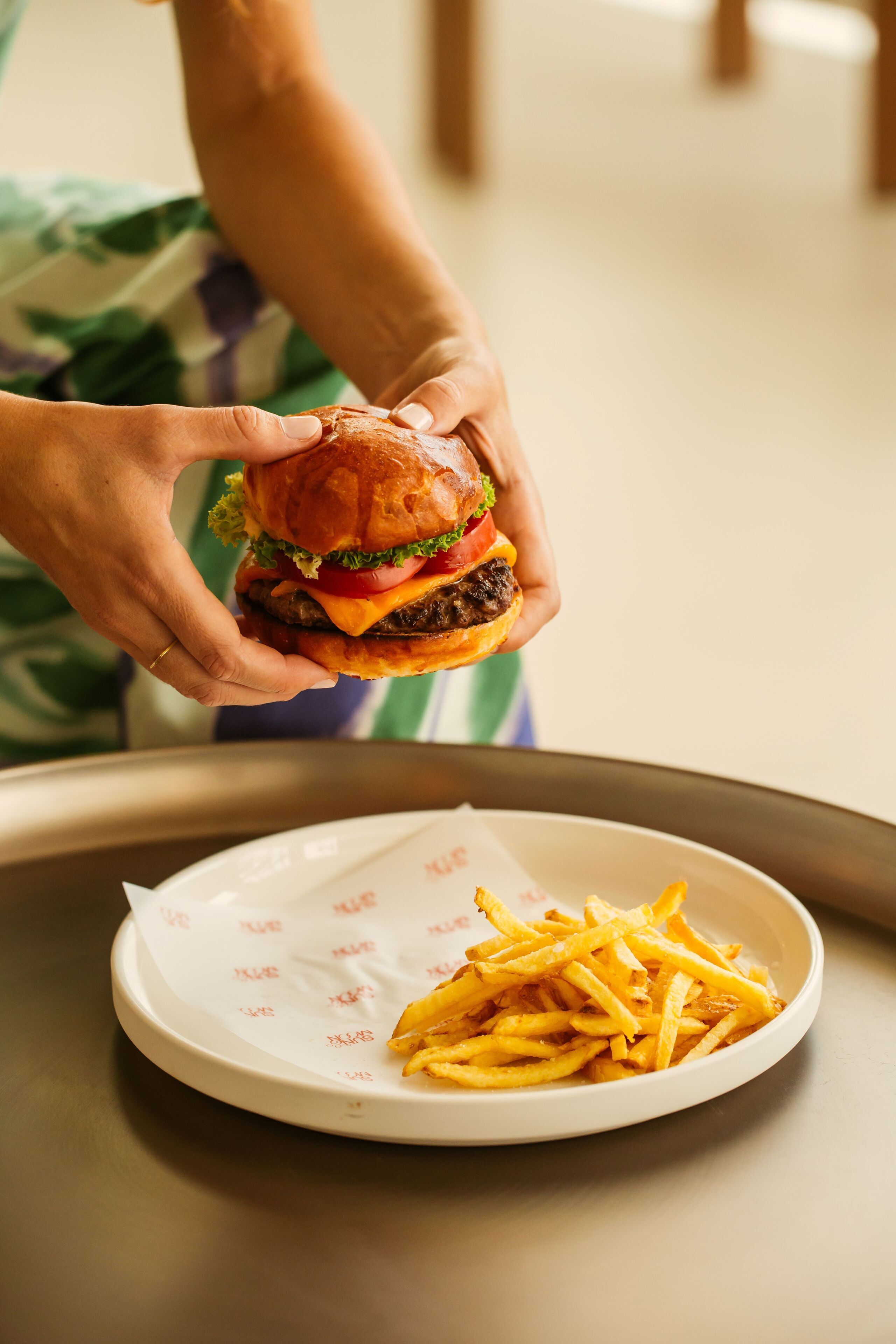A person holding a burger over a plate of fries.