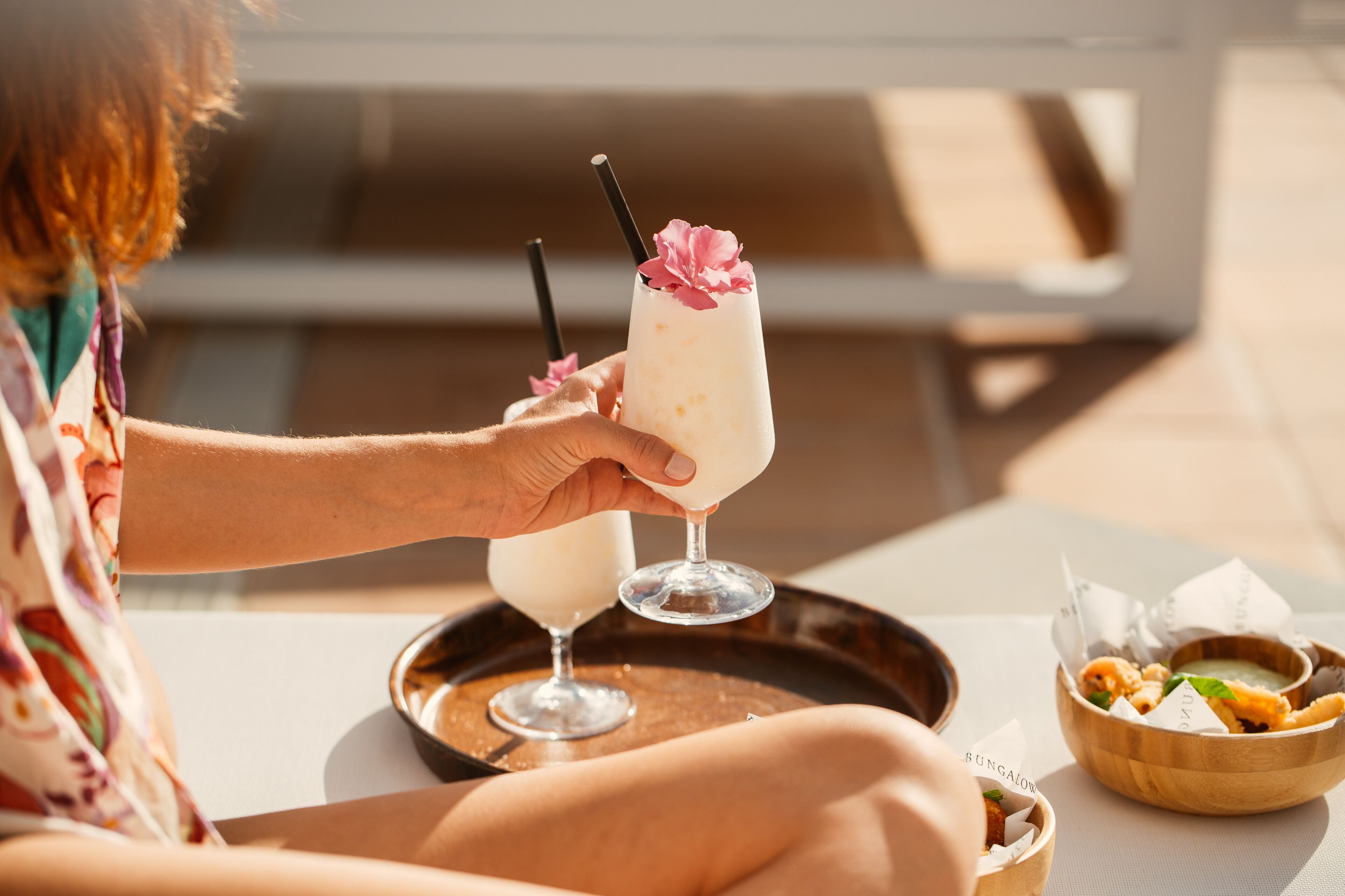 A person holding two drinks with straws, a tray with a bowl of food, and a wooden bowl with snacks.