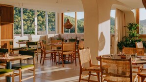 A spacious dining area with high ceilings, large windows offering a view of greenery, and a mix of wooden and woven furniture. - Mondrian Ibiza (Santa Eulalia del Rio)