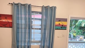 A living room with a brown sofa, a window with blue curtains, and two paintings on the wall. - Nutmeg Hill Villa (2 Bedrooms) (Soufriere)