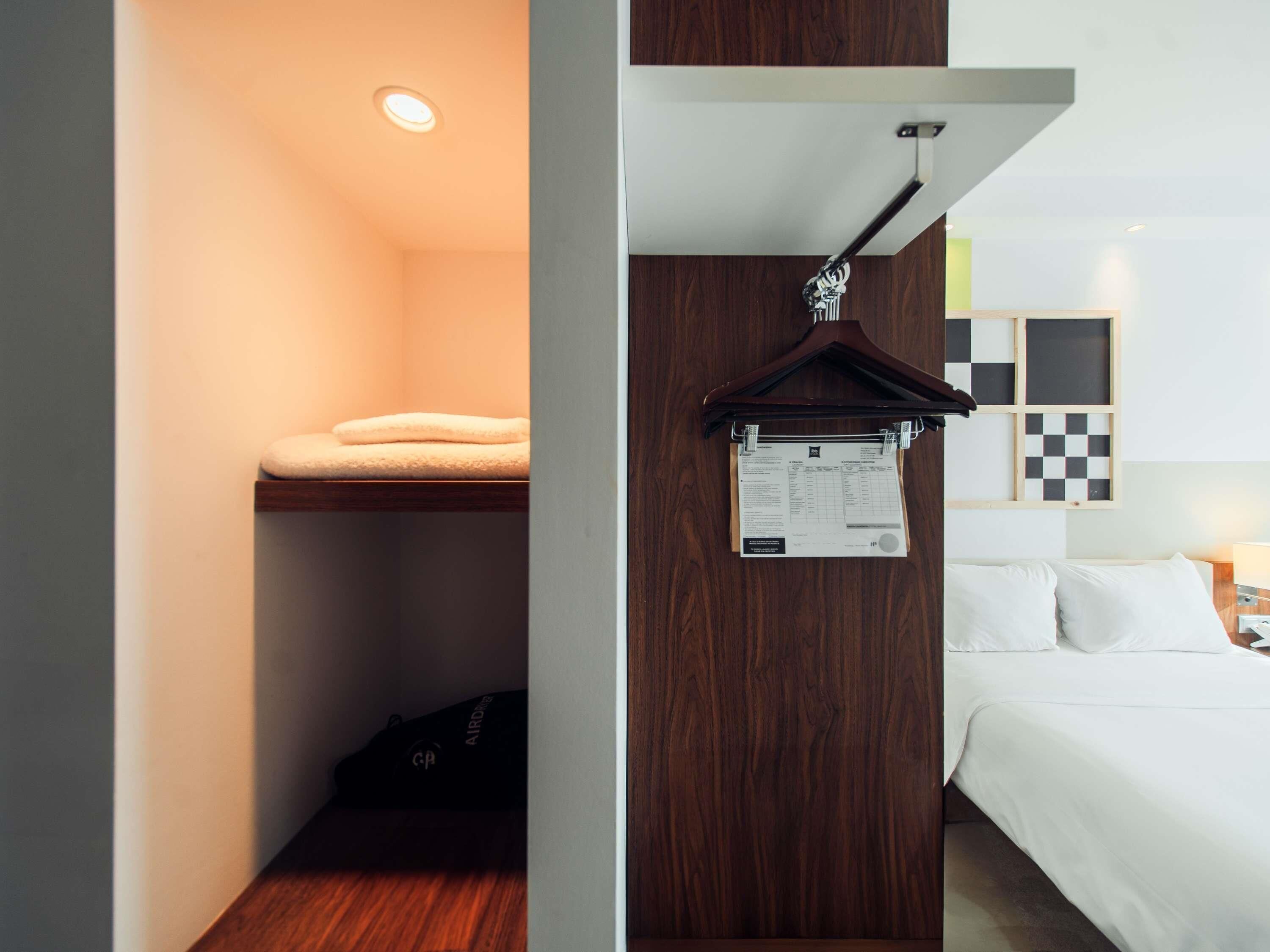 A hotel room with a bed, a hanging coat rack, a wooden wardrobe, and a small shelf with a folded towel.