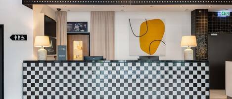 Una recepción de hotel con diseño de baldosas de cuadros, dos lámparas, una obra de arte en la pared y un cartel que indica instalaciones accesibles.