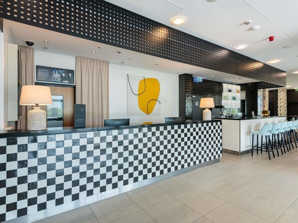 Zona de recepción de hotel con suelo a cuadros, mostrador de recepción, área de bar y un cuadro abstracto amarillo en la pared.