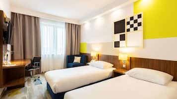 Habitación de hotel con dos camas, un escritorio, una silla, una lámpara de escritorio, un adorno de pared a cuadros y una ventana con cortinas.