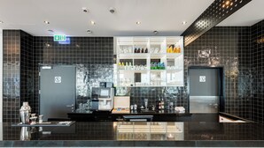 A modern bar area with a black-tiled backsplash, a glass display case with bottles, and a digital screen. - Ibis Styles Warszawa Airport (Warsaw)