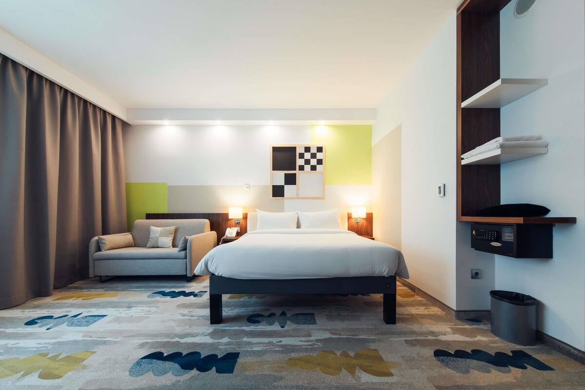 Ein modernes Hotelzimmer mit Bett, einer Sitzecke, einem Schreibtisch und einer integrierten Ablage mit einer Mikrowelle.