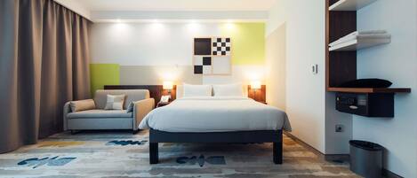 A modern hotel room with a bed, a sofa, a desk, and a built-in shelf with a microwave.