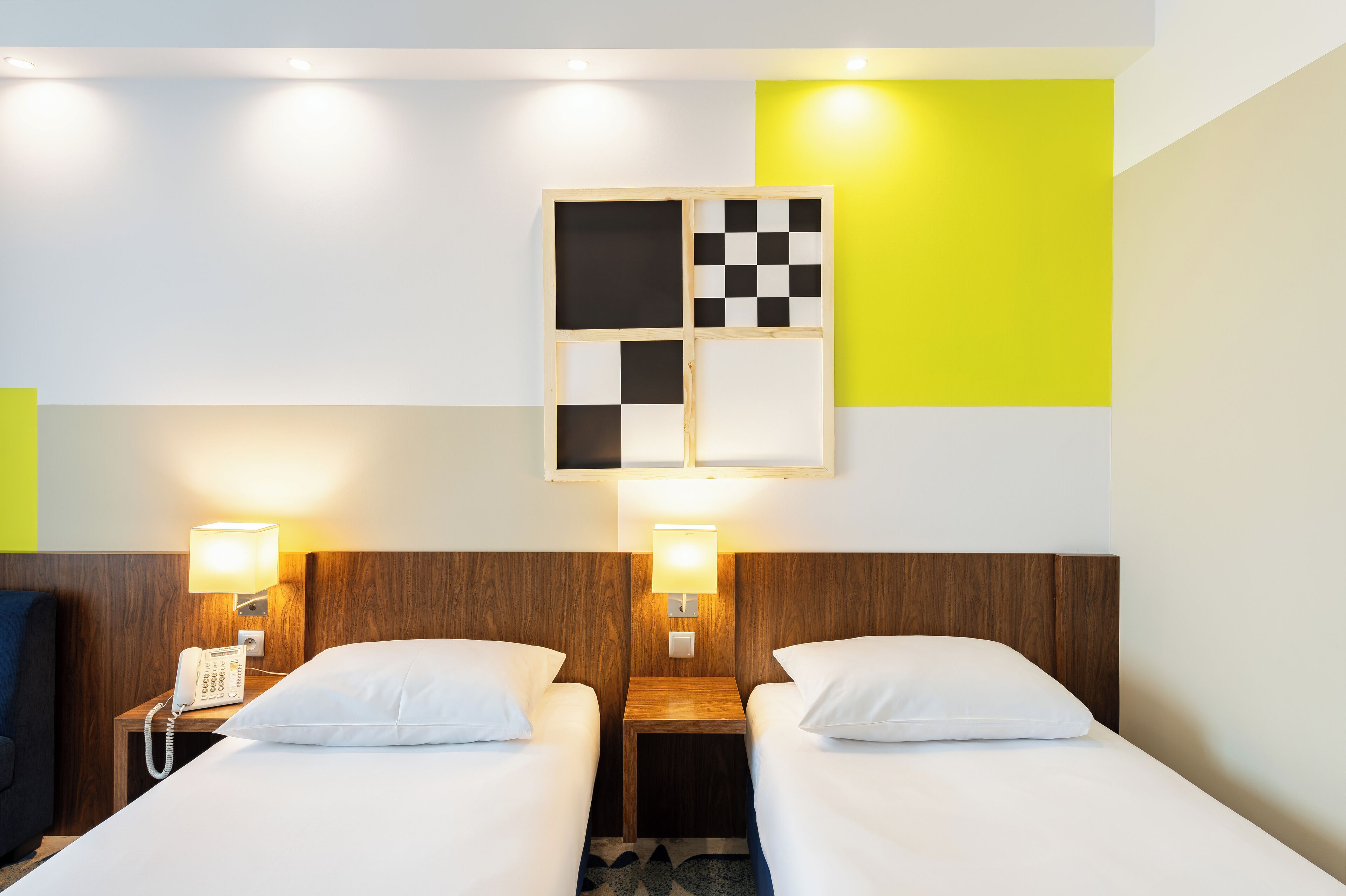 A hotel room with two beds, a wooden headboard, a nightstand with a phone, and a wall-mounted light fixture.