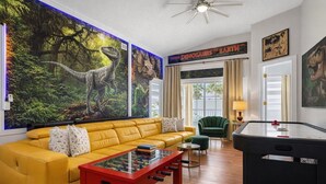 A room with a yellow sofa, a red coffee table, a foosball table, and a large mural of dinosaurs. - Animatronic Dinosaur Villa: 3 King Rooms, Theater & Private Pool Near Disney (Clermont)