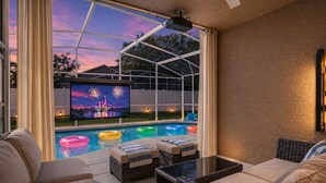 A pool area with a television displaying fireworks, outdoor seating, and a covered patio. - Animatronic Dinosaur Villa: 3 King Rooms, Theater & Private Pool Near Disney (Clermont)