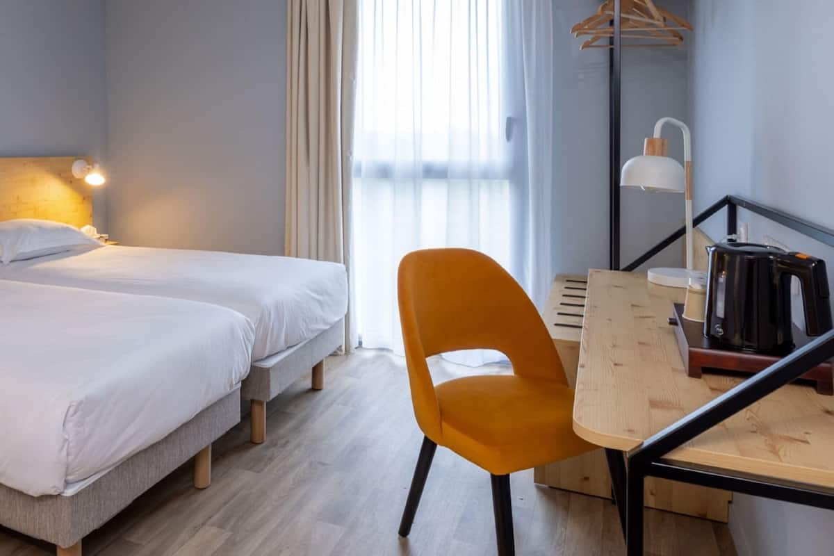 Une chambre d’hôtel avec deux lits, un bureau en bois, une chaise, une lampe et un porte-manteau mural.