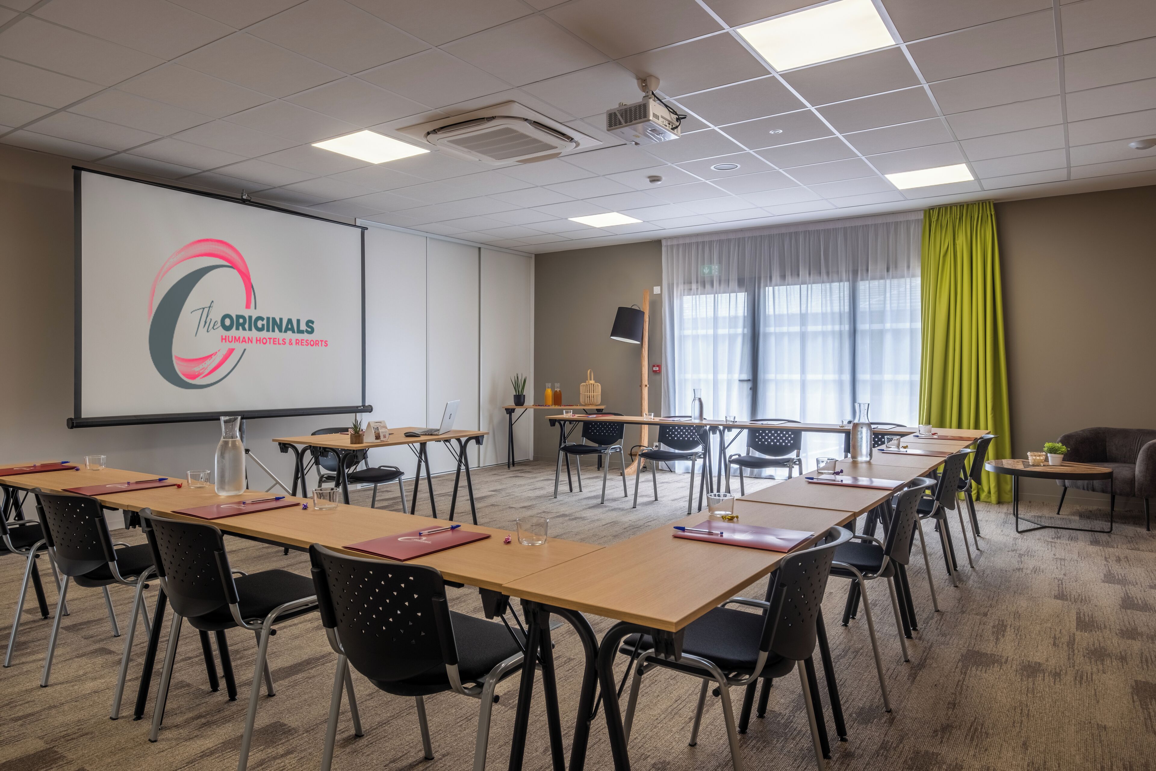 A conference room with a large screen displaying a logo, multiple tables with chairs, a projector, and a window with curtains.