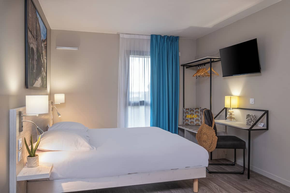 Une chambre d’hôtel comprenant un lit, une table de chevet, une lampe, une télévision, un bureau, une chaise et une fenêtre avec des rideaux.
