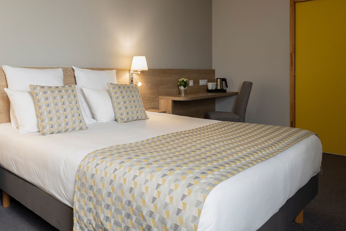 A hotel room with a bed, bedside table, lamp, and a small desk with a chair.