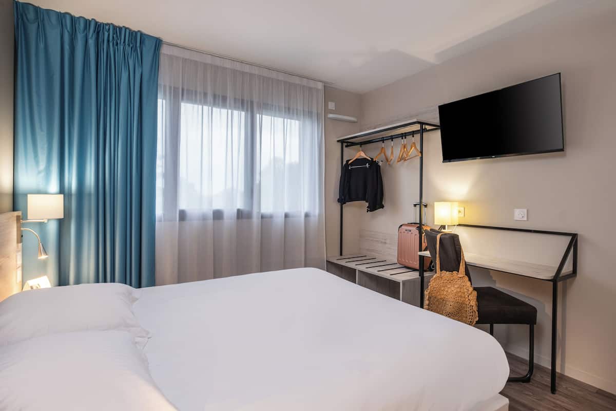 Une chambre d’hôtel avec un grand lit, un bureau avec une chaise, un téléviseur fixé au mur et un portant à vêtements avec des cintres.