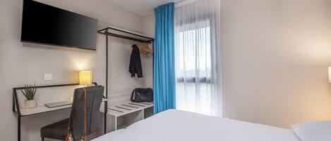 Quarto de hotel com uma cama, uma escrivaninha com cadeira, televisão na parede, luminária e janela com cortinas azuis.