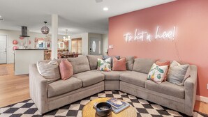 A modern living room with a sectional sofa, a ceiling fan, and a kitchen in the background. - Heated Deep Diving Pool, Firepit, BeautyBar, Yard Games, 3 King + 2 Queen beds (Tempe)