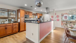 A modern kitchen with wooden cabinets, a central island, and a dining area with pink accents. - Heated Deep Diving Pool, Firepit, BeautyBar, Yard Games, 3 King + 2 Queen beds (Tempe)