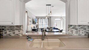 Een keuken met een groot eiland, een dubbele spoelbak en uitzicht op een woonkamer met een bank en een eethoek.