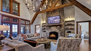 A spacious living room with a stone fireplace, wooden beams, and large windows.