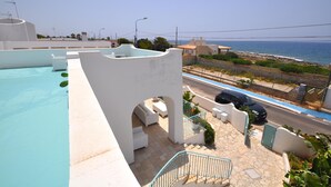 A rooftop pool area with a view of the sea, a car parked nearby, and a street in the background.