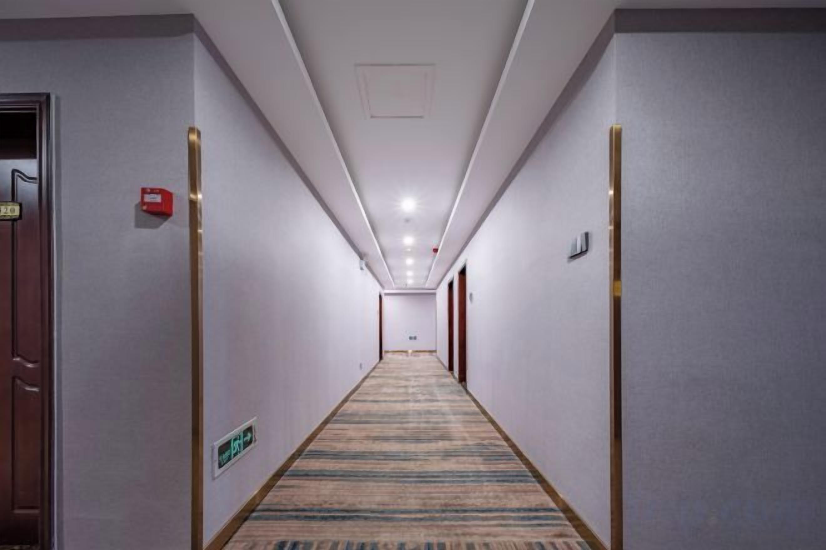 A hotel hallway with carpeted floors, doors on both sides, and a fire alarm on the left wall.