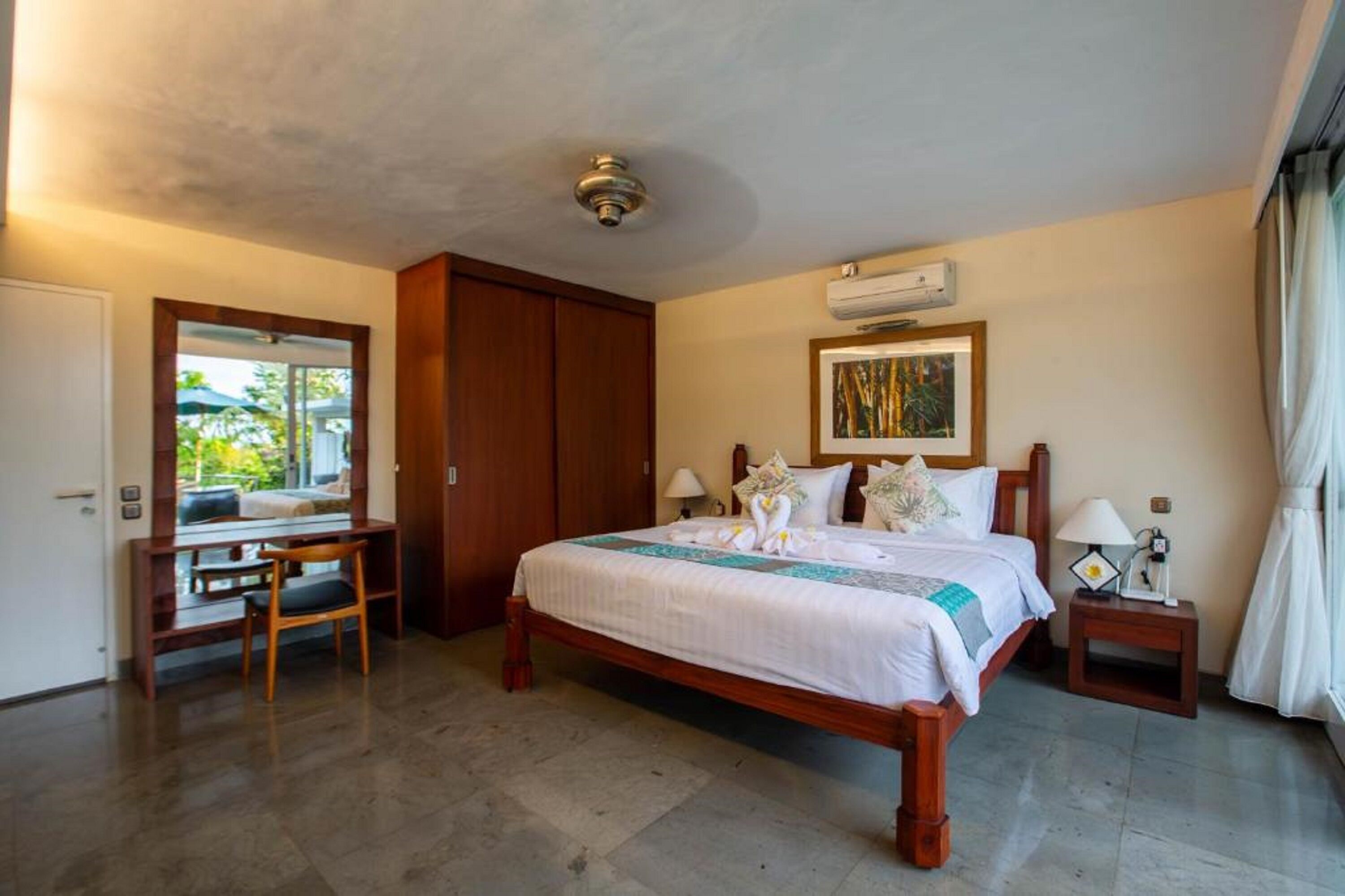 A hotel room with a wooden bed, a desk, and a chair. There is a window with a view of greenery outside.