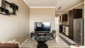 Una sala de estar moderna con una mesa de centro de vidrio, un televisor de pantalla plana y una zona de cocina con armarios oscuros y un refrigerador.