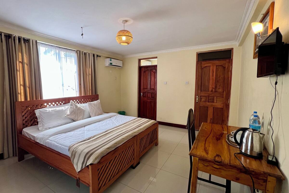 A hotel room with a wooden bed, a wooden desk with a kettle, a TV mounted on the wall, and a window with curtains.