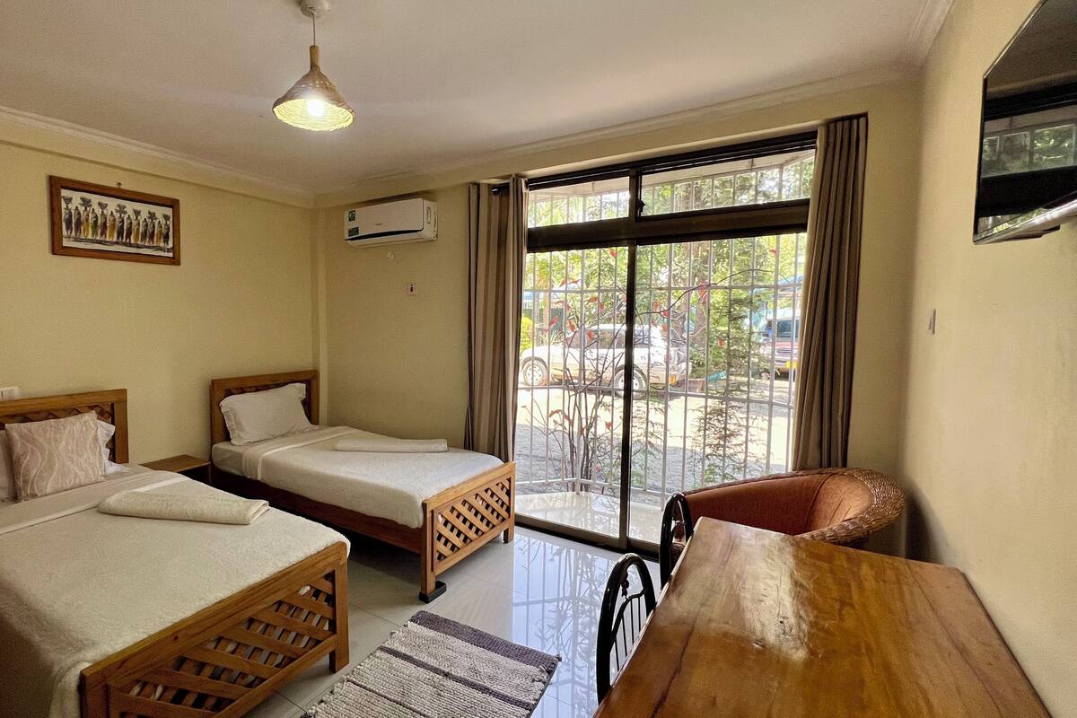 A room with two single beds, a wooden table, a chair, a ceiling light, a window with curtains, and an air conditioning unit.