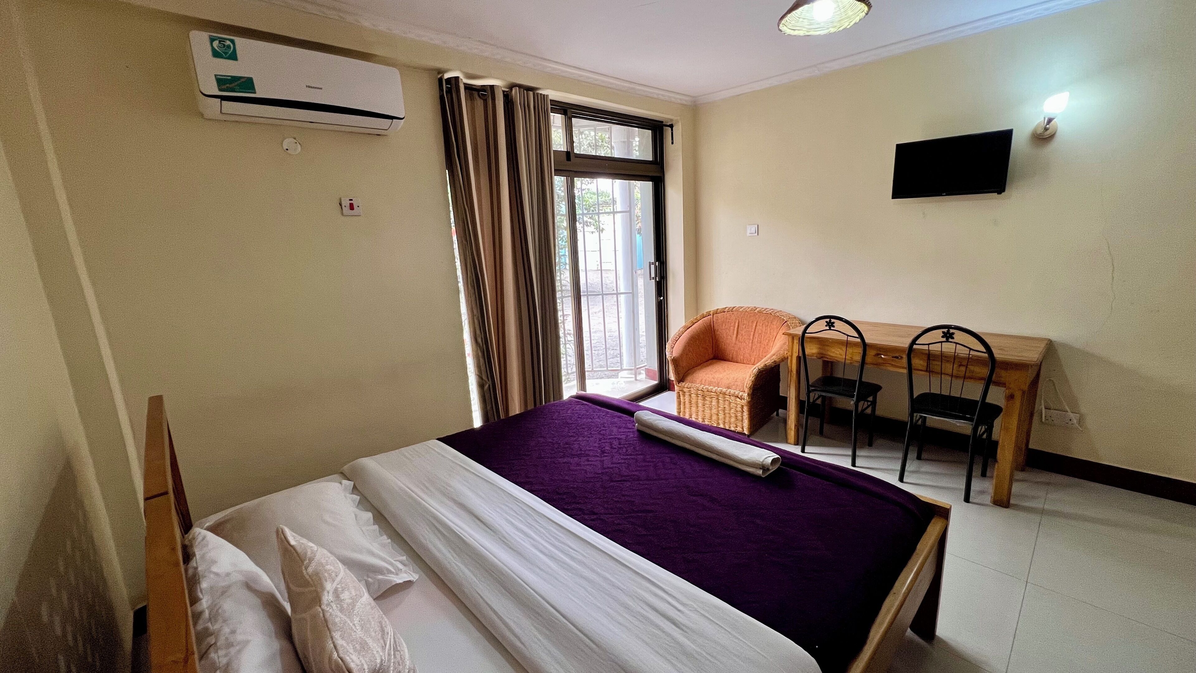 A bedroom with a bed, a desk, two chairs, a television, and an air conditioning unit.