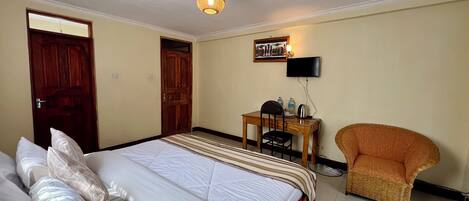 A hotel room with a bed, a desk with a chair, a television, and a wicker armchair.