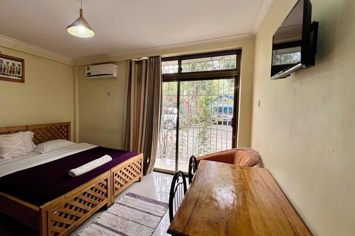 A hotel room with a bed, a wooden desk, a window with curtains, an air conditioning unit, and a television mounted on the wall.