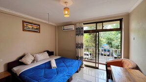 A bedroom with a bed, a nightstand, a chair, a window with curtains, and an air conditioning unit. - Runako Lodge (Arusha)