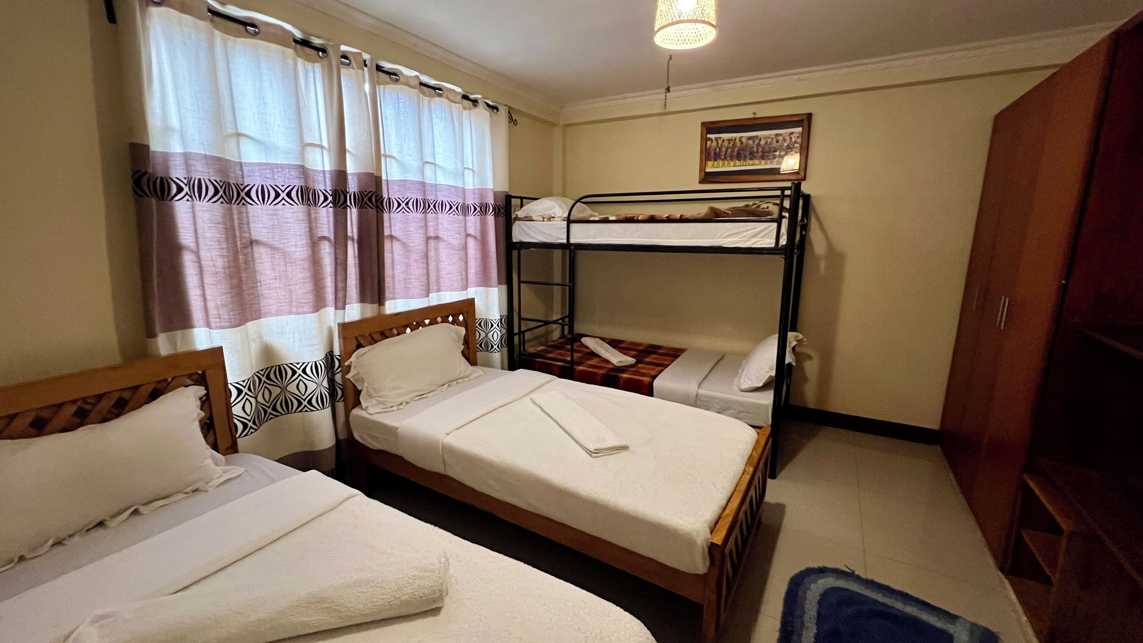 Habitació d'hotel amb dos llits individuals, una lliterà i un armari de fusta, un quadre amb marc a la paret i una finestra amb cortines.