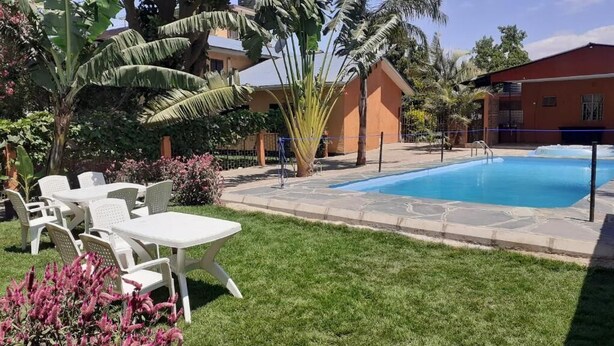 A pool area with a clear blue pool, surrounded by a stone deck, and adjacent to a building with a red roof. There are white plastic chairs and a small table on the grass. - Runako Lodge (Arusha)