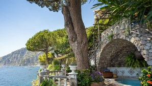 Un puente de piedra que conduce a una zona de piscina con vista al mar y un gran árbol.