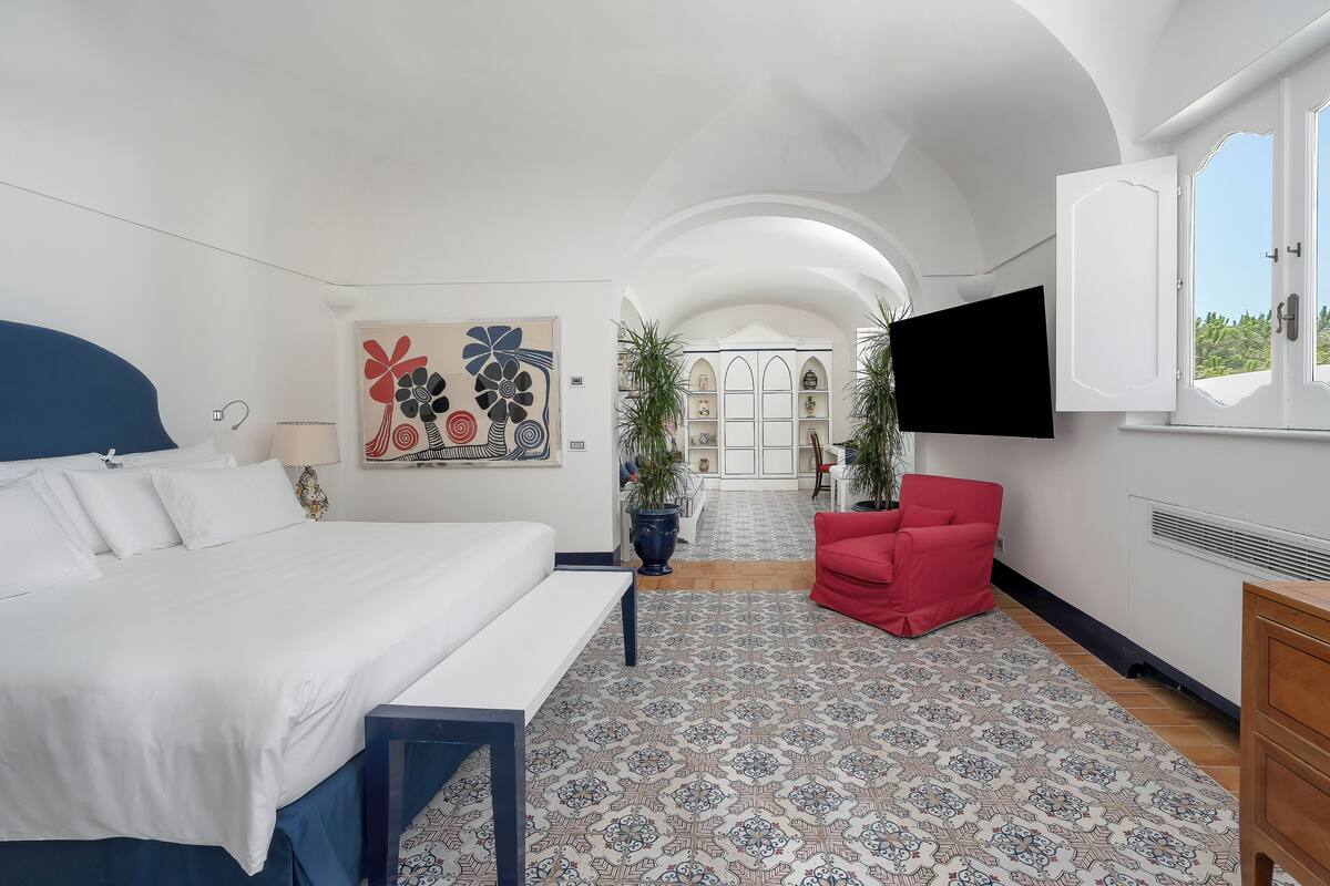 Un dormitorio con una cama, una silla roja, un televisor y vistas a un pasillo con una puerta.