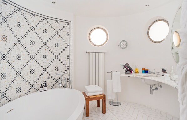 A modern bathroom with a freestanding bathtub, a wooden stool, a white sink with various toiletries, and a patterned wall. - Treville Positano (Positano)