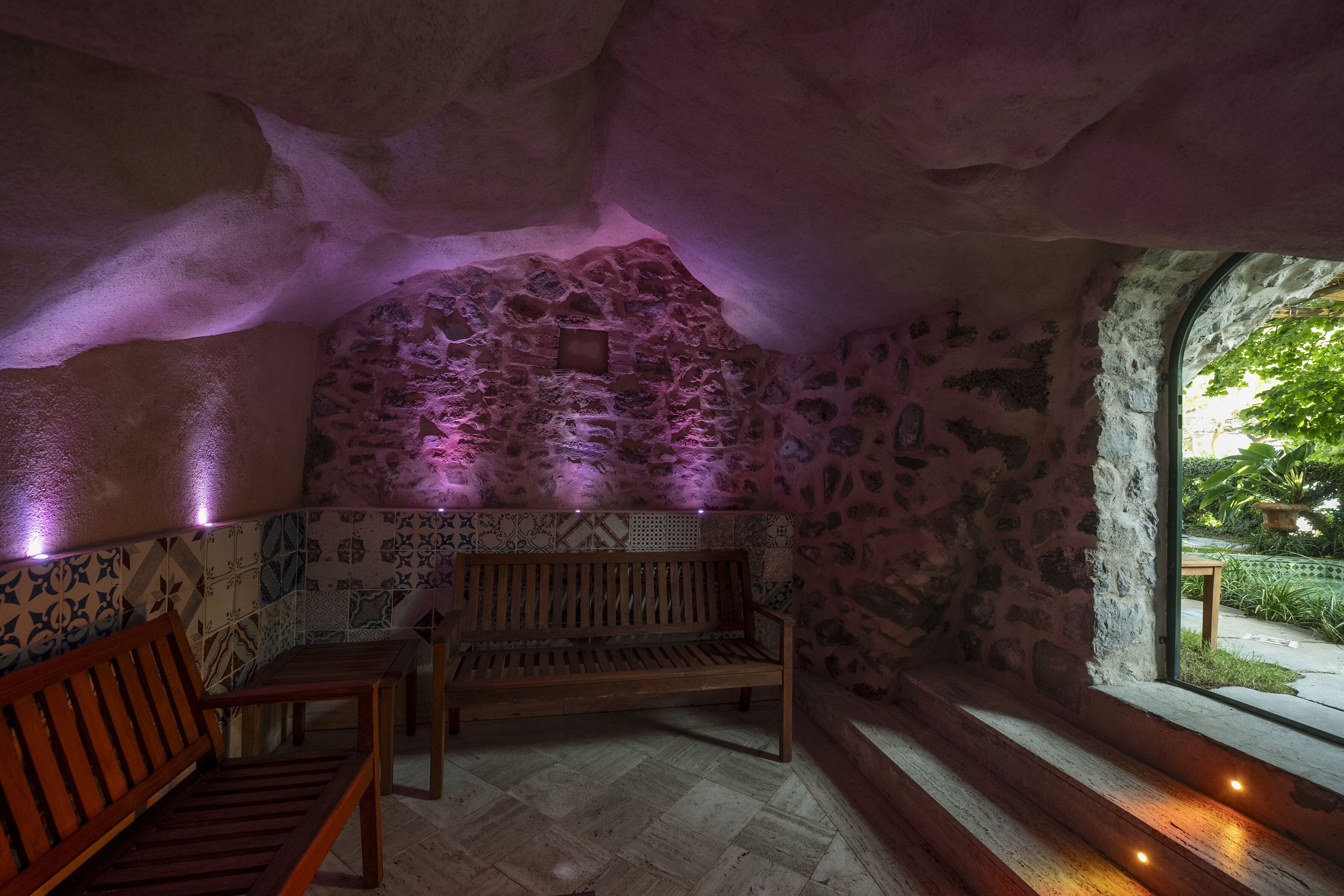 A stone-walled room with a wooden bench and a table, a window with a view of greenery, and purple lighting.