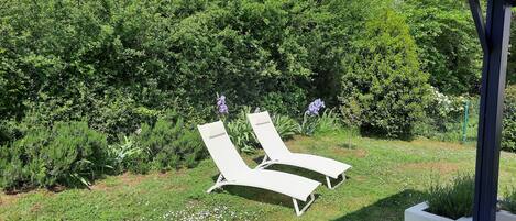 Two white lounge chairs in a garden with dense greenery and a clear sky.