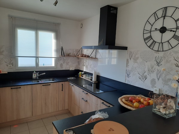 A modern kitchen with a black countertop, wooden cabinets, a large wall clock, and a window with blinds. - A charming, well-equipped house just a 5-minute walk from two beautiful beaches and the water sports center. (Pénestin)