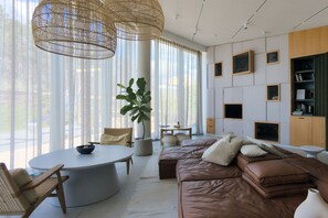 En moderne stue med en brun lædersofa, et rundt hvidt bord, pendellamper i flet og store vinduer med lette gardiner.