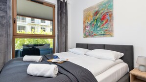 A neatly made bed with a dark quilt, white pillows, and a blue blanket. There is a window with a view of a building and greenery outside. - Gdańska 21R /5 (Gdansk)