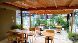 A covered outdoor seating area with wooden tables and chairs, potted plants, and a view of the street and greenery outside. - Casablanca (Santiago de Surco)