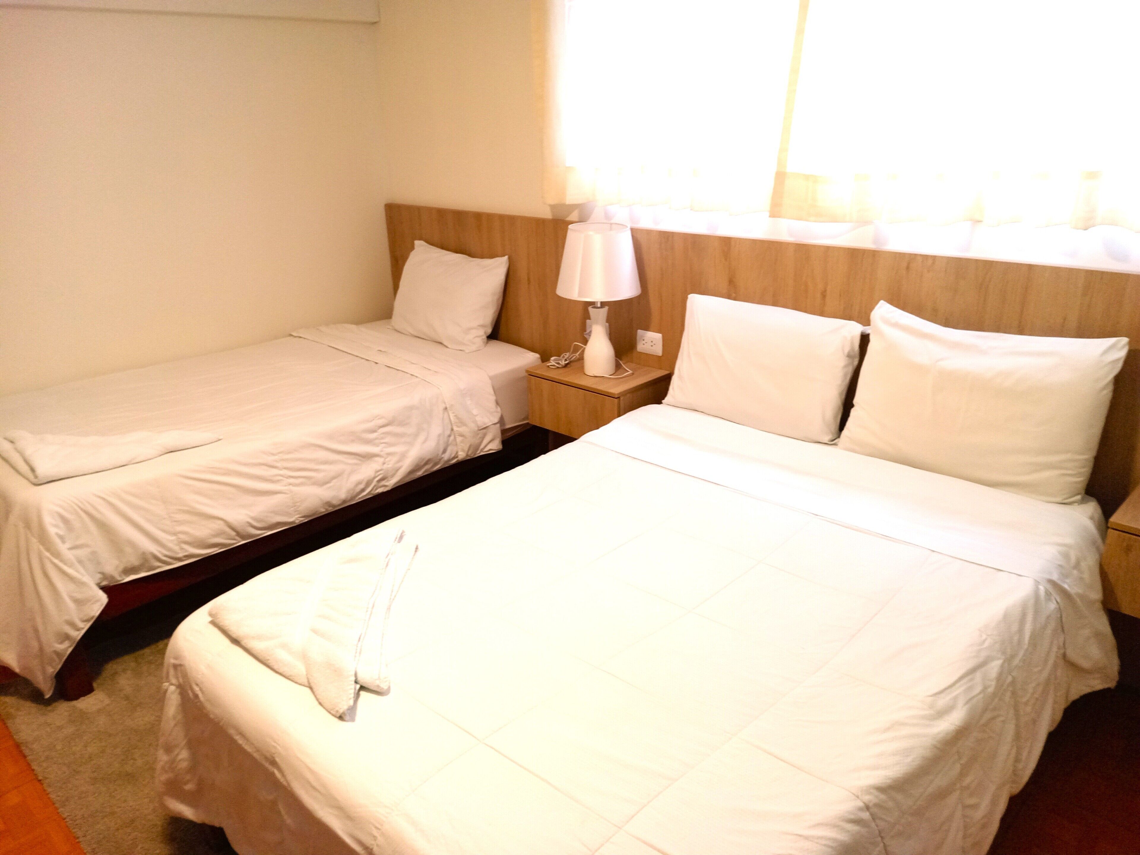 A hotel room with two single beds, a nightstand with a lamp, and a window with curtains.